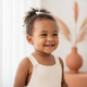 Joyful toddler smiling, headshot portrait against a soft warm amber and white background with terracotta accents.